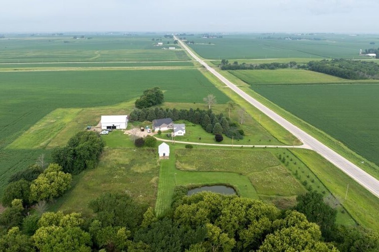 Aerial view of a rural Iowa acreage surrounded by farmland, with a home, outbuildings, driveway, pond and nearby roadway.
